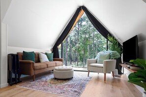 A modern loft bedroom with a sloped ceiling, a large window overlooking a forest, a brown sofa, a white armchair, and a flat-screen TV. - Coldwater Cove - New Zook Cabin on 19 Acres (Elberton)