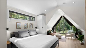 A modern bedroom with a large bed, a skylight, a lofted seating area with a view of trees, and artwork on the walls.