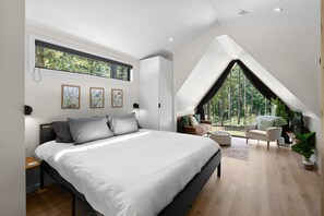 A modern bedroom with a large bed, a skylight, a lofted seating area with a view of trees, and artwork on the walls.