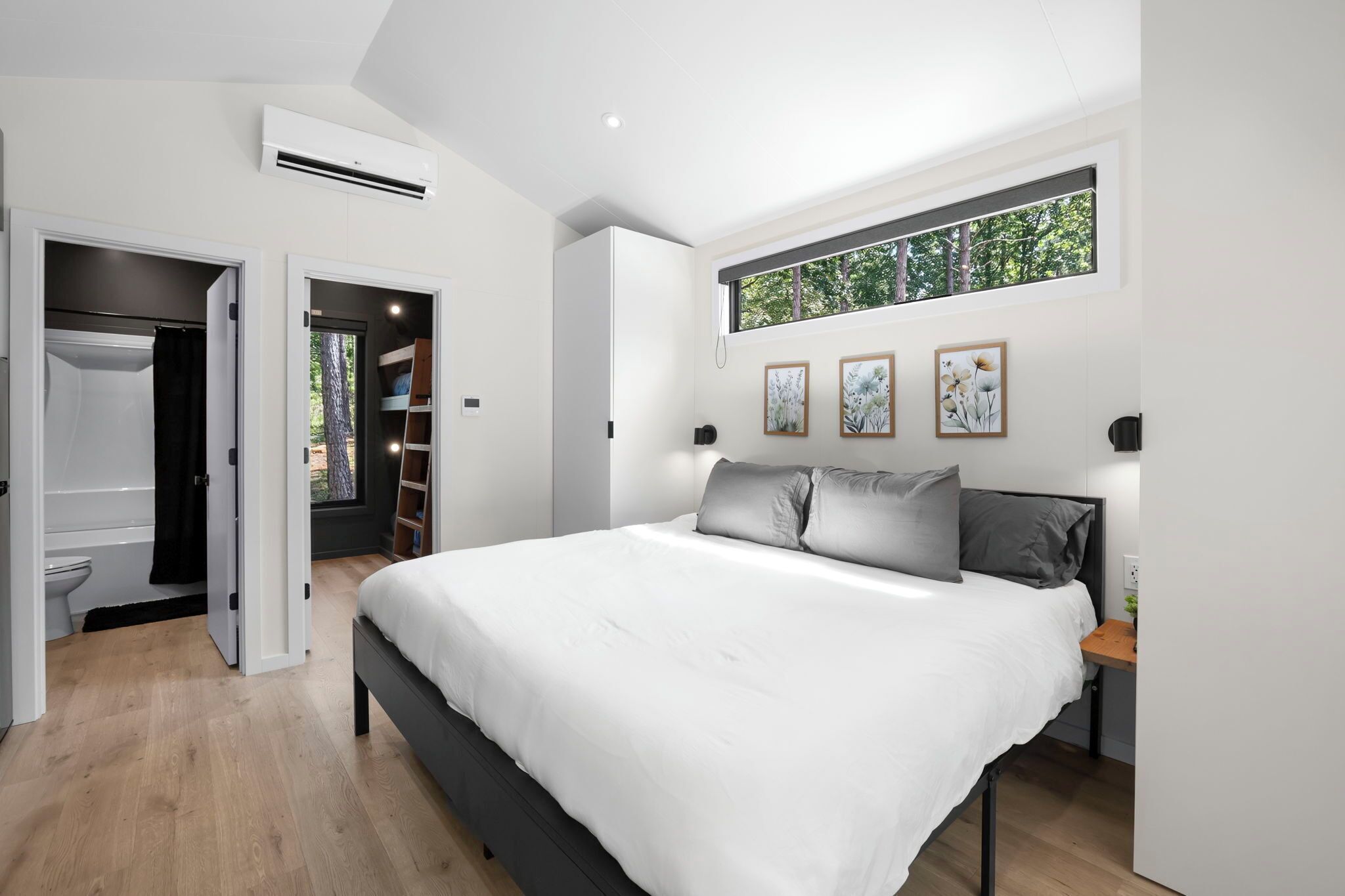 A modern bedroom with a large bed, a wardrobe, a built-in bench, and a window with a view of trees.