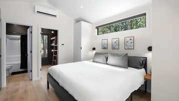 A modern bedroom with a large bed, a wardrobe, a built-in bench, and a window with a view of trees.