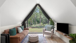 A modern, triangular room with a sofa, ottoman, and armchair, a flat-screen TV, and a large window overlooking a forest.