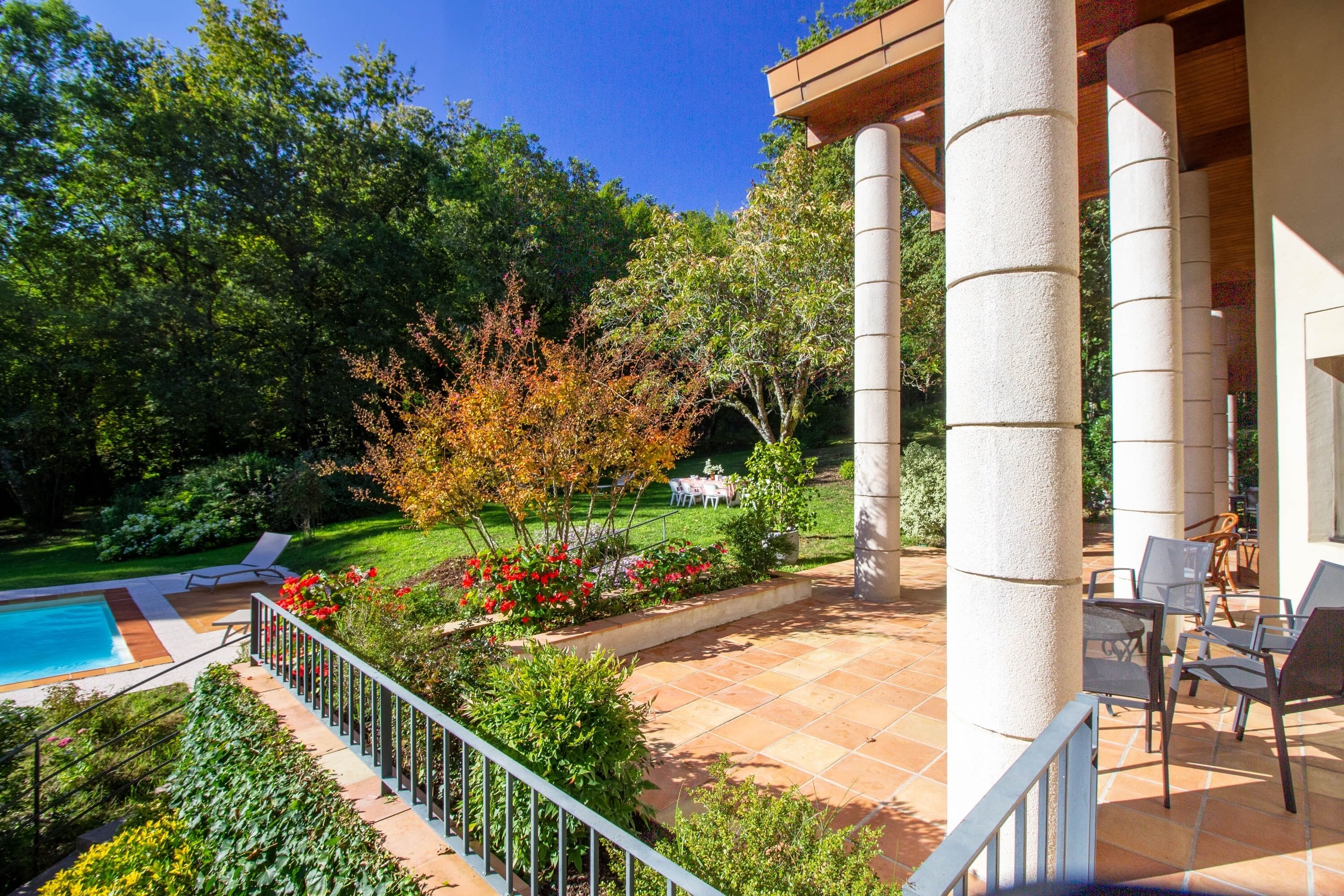 Zona de piscina con terraza, asientos y vistas a un jardín y árboles.