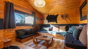 Ein gemütliches Blockhaus-Interieur mit Holzbalkendecke, einem großen Fenster, einer Couch, einem Couchtisch und einem aufgehängten Tierfell.