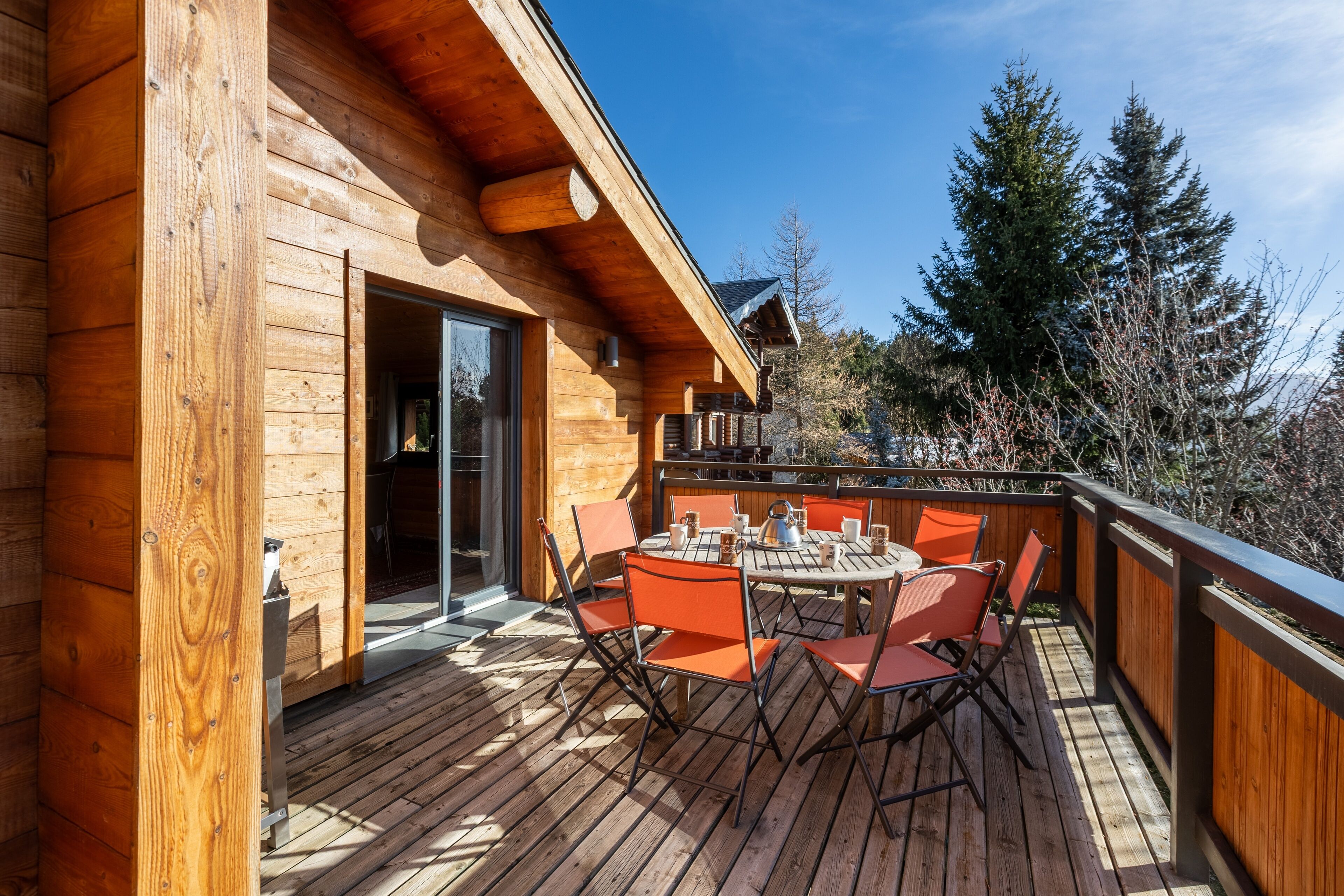 Une terrasse en bois avec une table ronde et des chaises orange, une porte d’entrée en verre et une vue sur les arbres.