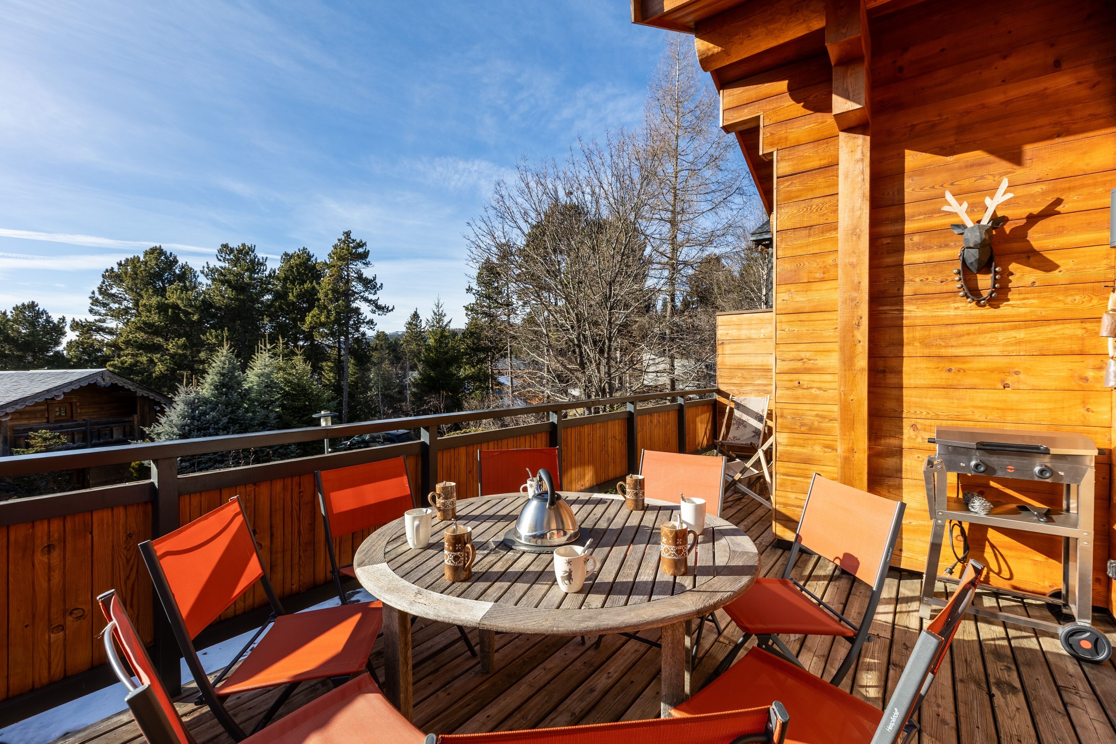 A wooden deck with a round table and chairs, a kettle, mugs, and a grill.