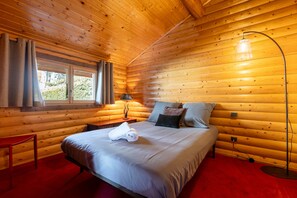 Ein Zimmer in einer Holzhütte mit einem Bett, einem Nachttisch, einer Lampe und einem Fenster mit Vorhängen.