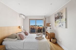 A bedroom with a bed, pillows, a nightstand, a bedside table, a wall-mounted air conditioner, a large window, and a view of the outdoors. - Beaulieu Palace - Rooftop Vue Mer (Beaulieu-sur-Mer)