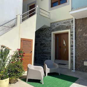 A building with a stone facade, a wooden door, a balcony, and two plastic chairs on a green surface.
