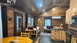 Interior acogedor de una cabaña con una mesa de comedor de madera, un sofá, un árbol de Navidad y una zona de cocina.