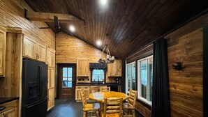 Interior de una cabaña de madera con una mesa de comedor, sillas, un refrigerador y una zona de cocina.