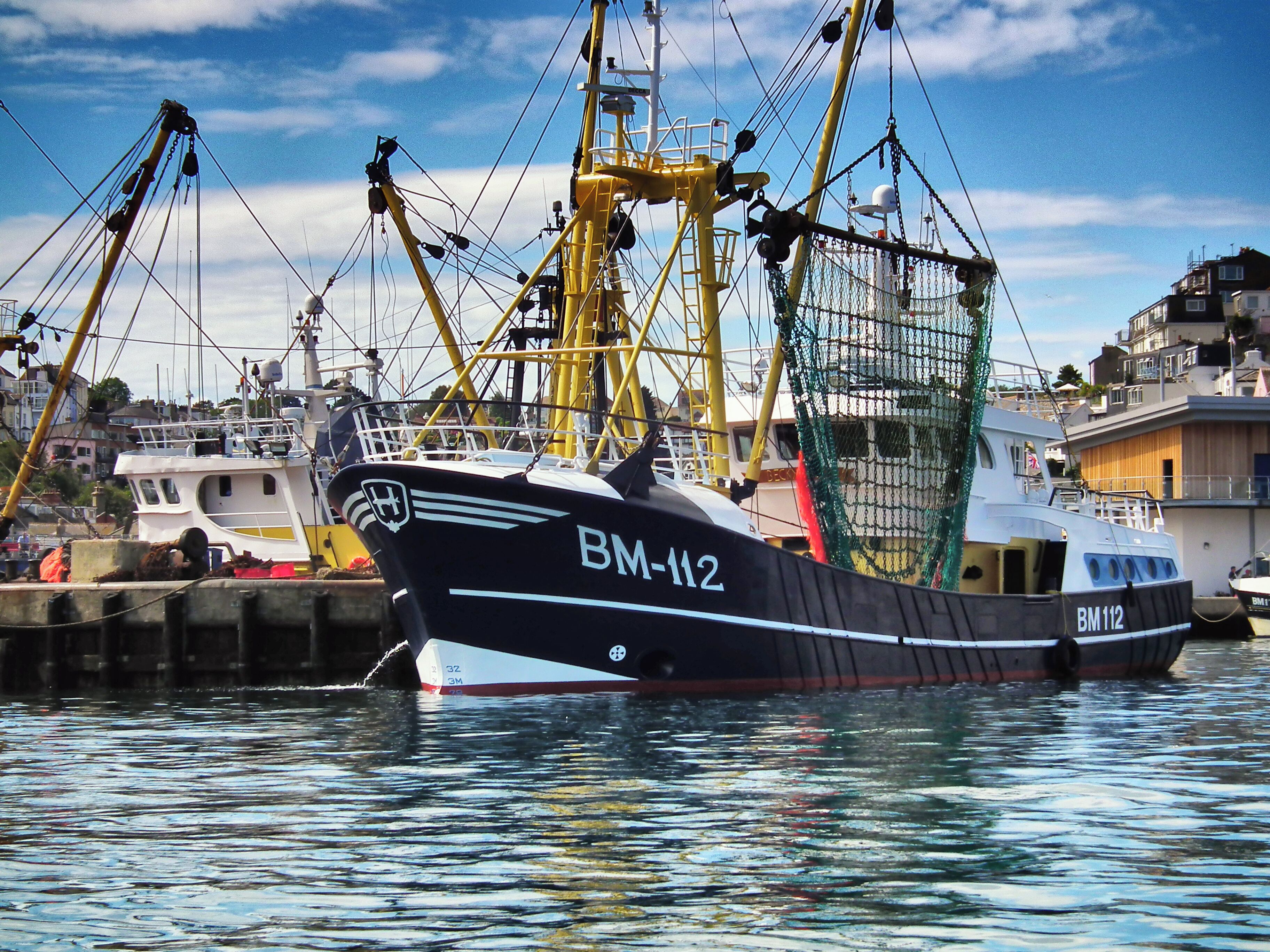 Ein Fischerboot mit dem Namen BM-N2 liegt im Hafen vor dem Hintergrund von Kränen und Gebäuden.