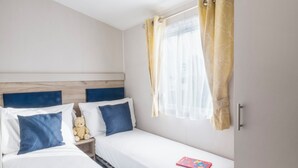 A bedroom with two beds, a nightstand, a teddy bear, and a window with curtains. - Caravan C32 at Landscove Holiday Park (Brixham)