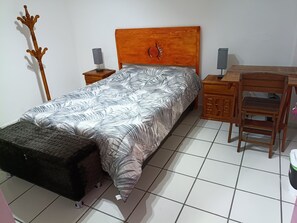 A bedroom with a bed, a wooden headboard, a nightstand with a lamp, a desk with a chair, and a wooden coat rack.