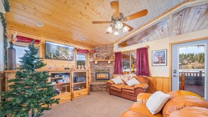 A cozy cabin interior with a stone fireplace, wooden furniture, a Christmas tree, and a large window overlooking a scenic landscape. - Hot Tub with Meadow Views! Pet Friendly! (Duck Creek Village)