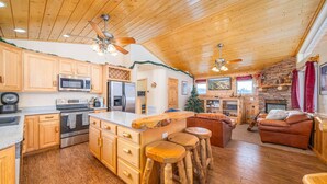 A spacious kitchen with wooden cabinets, a central island with stools, a stone fireplace, and a cozy seating area with a leather couch. - Hot Tub with Meadow Views! Pet Friendly! (Duck Creek Village)