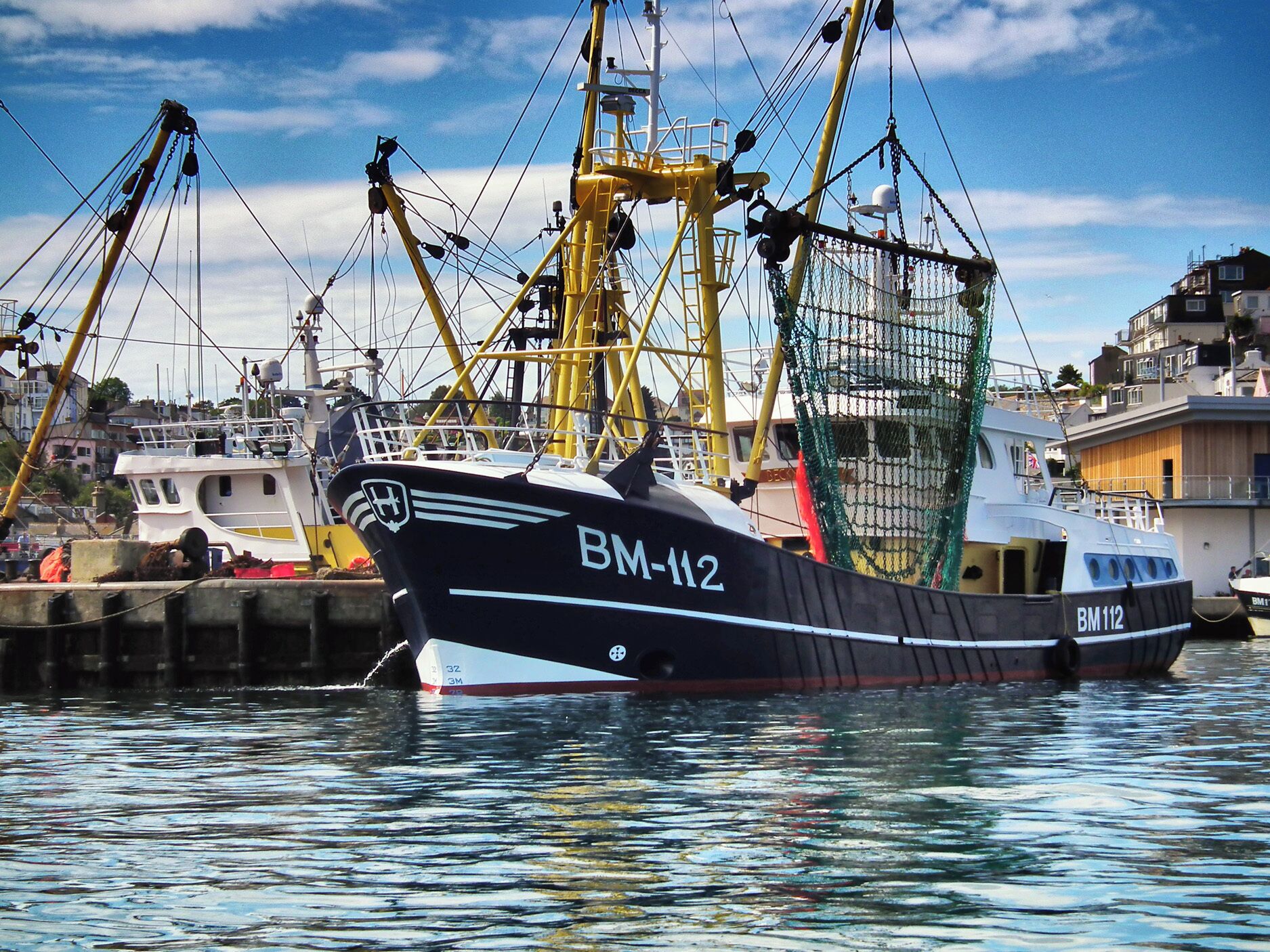 Ein Fischerboot mit dem Namen BM-112 liegt im Hafen festgemacht, im Hintergrund sind ein Kran und weitere Boote zu sehen.