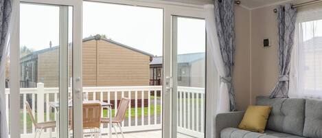A room with a sliding glass door leading to an outdoor deck, a grey sofa with a yellow pillow, and a small table with chairs.