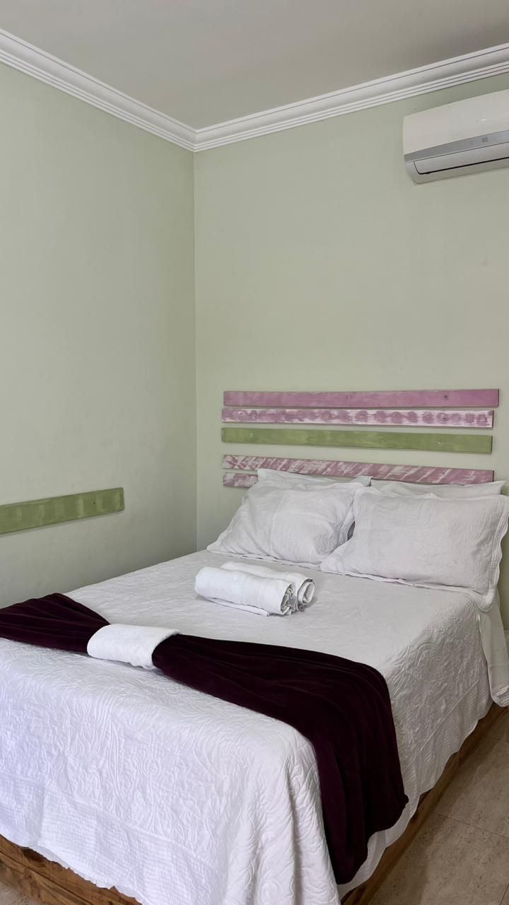 Una cama bien hecha con sábanas blancas y una manta roja oscuro, un cabecero de madera con paneles decorativos y un aire acondicionado instalado en la pared.