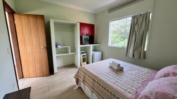 Un dormitorio con cama, armario, televisor sobre una estantería y ventana con cortinas.