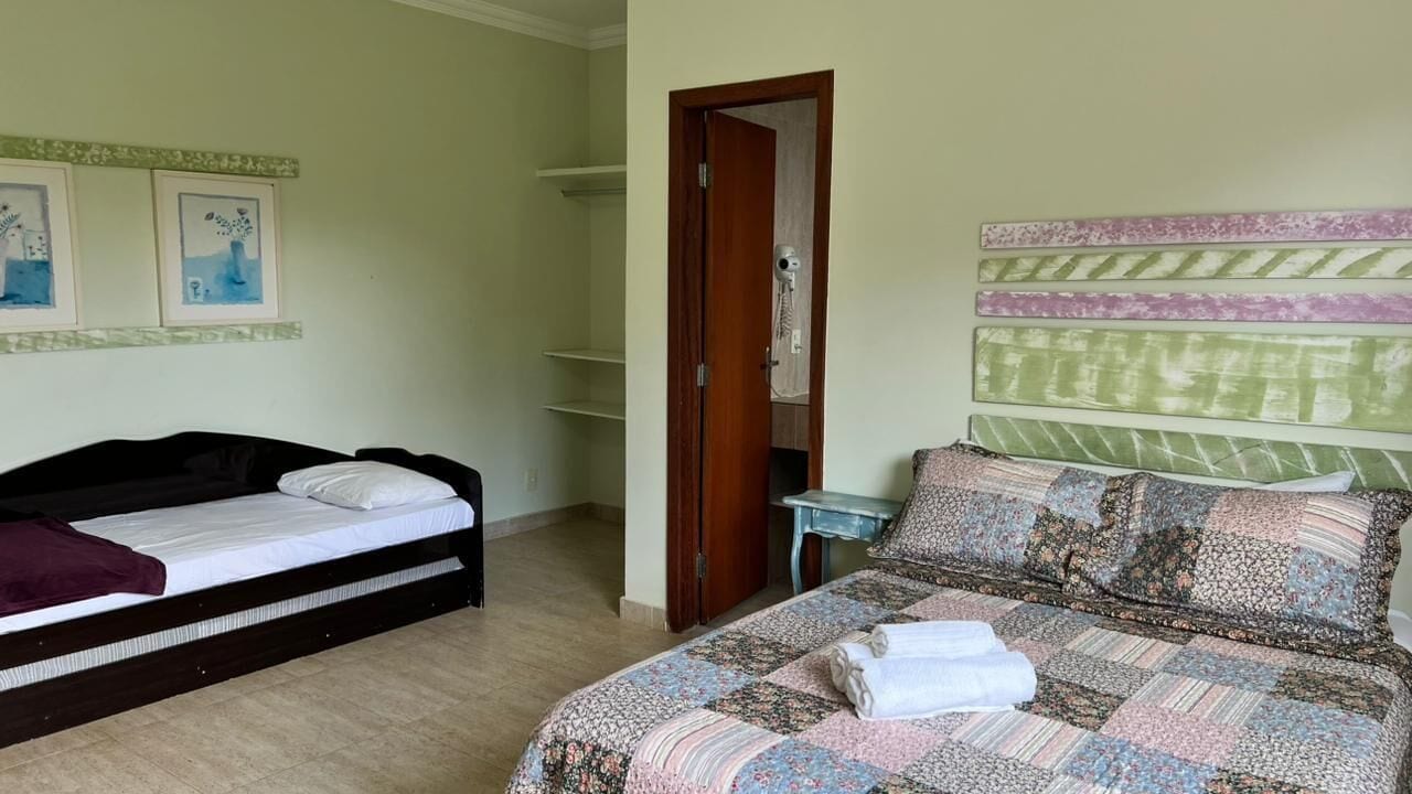 A bedroom with a bed on the left and a larger bed on the right, both with pillows and blankets. There is a wooden door leading to a bathroom, and a small table with a lamp next to the larger bed.