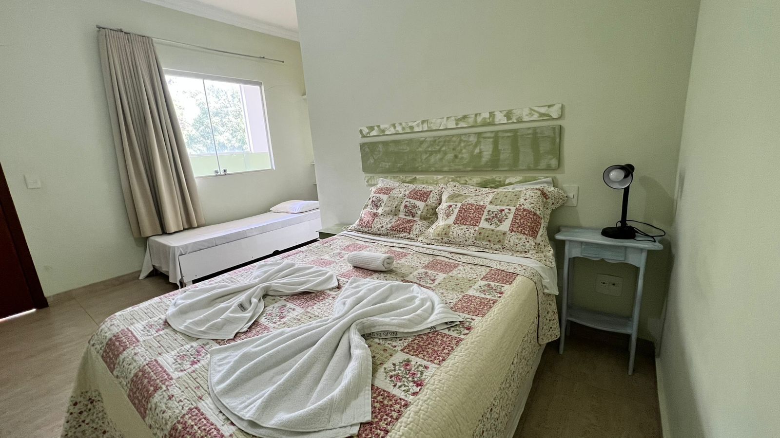 A neatly made bed with a floral-patterned bedspread and pillows, a bedside table with a lamp, and a window with curtains.
