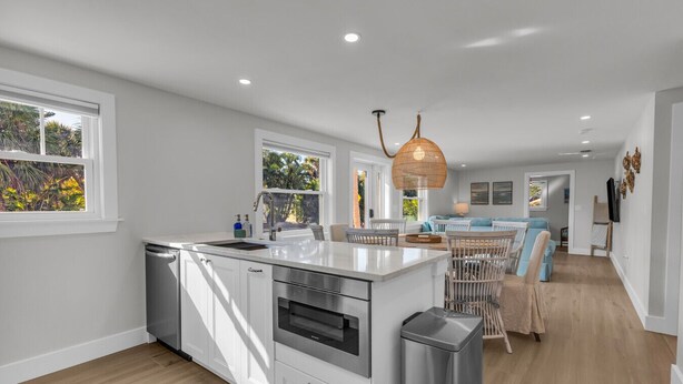 A modern kitchen with white cabinetry, a built-in oven, and a large island with a sink. There is a dining area with a blue sofa and wicker chairs. - The Pink House - Walking Distance to the Beach and Restaurants (Holmes Beach)