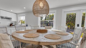 A round wooden dining table with woven placemats, surrounded by white chairs, in a bright kitchen with white cabinets and a large window. - The Pink House - Walking Distance to the Beach and Restaurants (Holmes Beach)