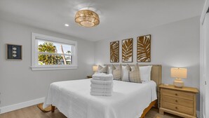 A neatly arranged bedroom with a large bed, a bedside table with a lamp, and a window with a view of palm trees. - The Pink House - Walking Distance to the Beach and Restaurants (Holmes Beach)