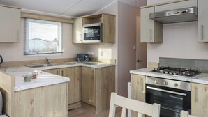 A compact kitchen with wooden cabinets, a built-in microwave, a gas stove, and a sink. - Caravan B61 at Landscove Holiday Park (Brixham)