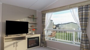 A living room with a flat-screen TV, a small shelf with decorative items, a window with a view of a house, and a balcony with a white railing. - Caravan B61 at Landscove Holiday Park (Brixham)