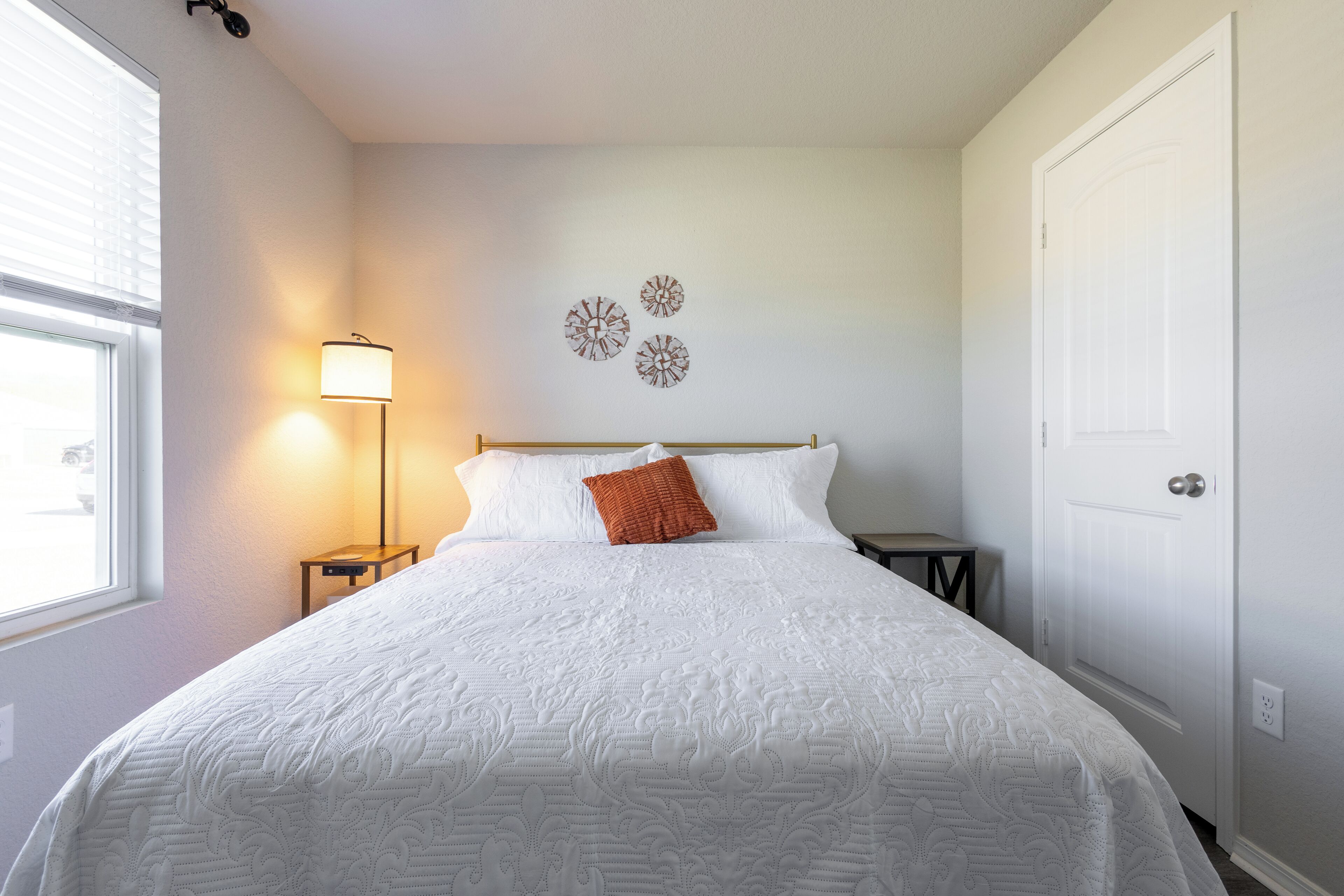 Een keurig opgemaakt bed met een witte quilt, één oranje kussen en een bedlampje.