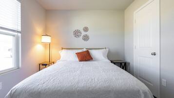 Een keurig opgemaakt bed met een witte quilt, één oranje kussen en een bedlampje.