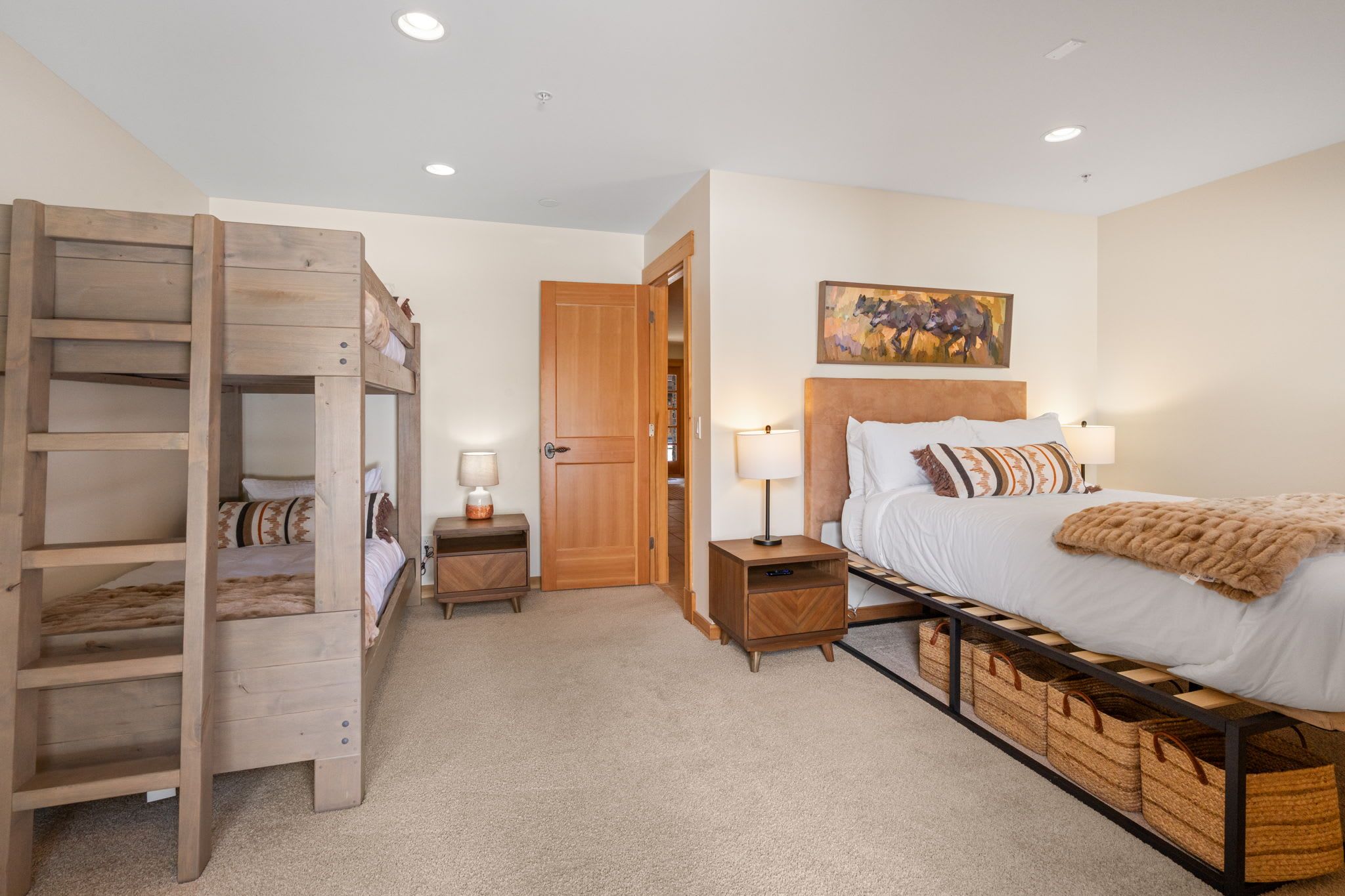 A bedroom with a bunk bed, a single bed with a headboard, bedside lamps, a painting on the wall, and storage baskets under the bed.