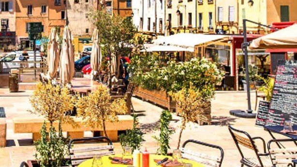 Un café en plein air avec des tables et des chaises rondes, entouré de bâtiments et de verdure.