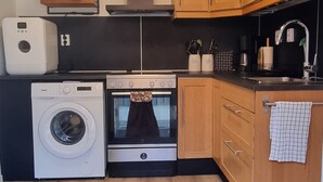 A compact kitchen with a washing machine, oven, microwave, and wooden cabinets. - Lyckeby Lodge – A Quiet Apartment Near Nature, Sea & City (Lyckeby)