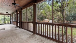 Une véranda couverte avec un plancher et des balustrades en bois, de grandes fenêtres offrant une vue sur les arbres environnants.