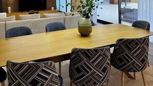 Een moderne eetruimte met een lange tafel, gestreept beklede stoelen, een vaas met groen en een televisie aan de muur.