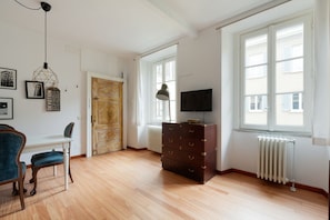 Ein Zimmer mit Holzboden, einem Schreibtisch mit Stuhl, einem Schminktisch, einem Fernseher und einem Fenster mit Jalousien.