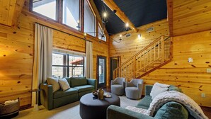 A cozy log cabin interior with a staircase, two green sofas, a round ottoman, and a large window. - Waterfront Cabin • Pond • Hot Tub • Great Location (Broken Bow)