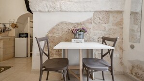 Una habitación con pared de piedra, una mesa blanca con dos sillas, una pequeña cocina y un letrero que dice 'Bienvenue'.