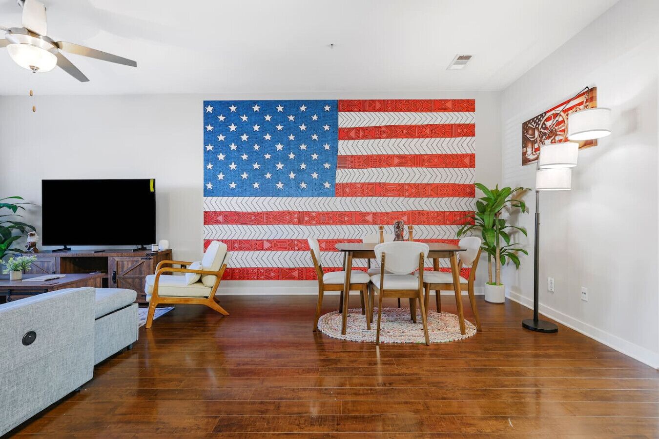A living room with a large American flag mural, a flat-screen TV, a dining table with chairs, and a floor lamp.