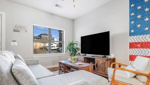 Ein Wohnzimmer mit grauem Sofa, einem Holztisch, einem Fernseher und einem Fenster mit Blick auf Gebäude.