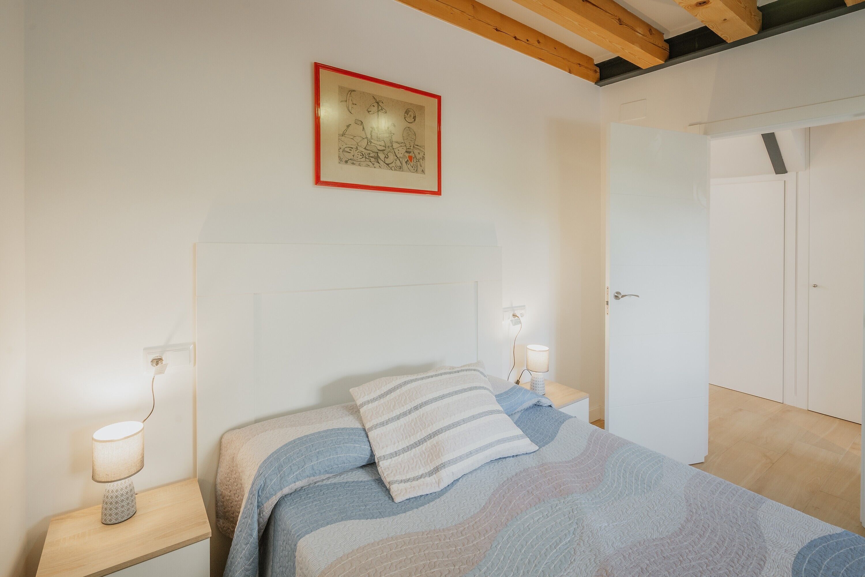 Une chambre à coucher avec un lit, des lampes de chevet, un tableau encadré au mur et une porte sur le côté droit.