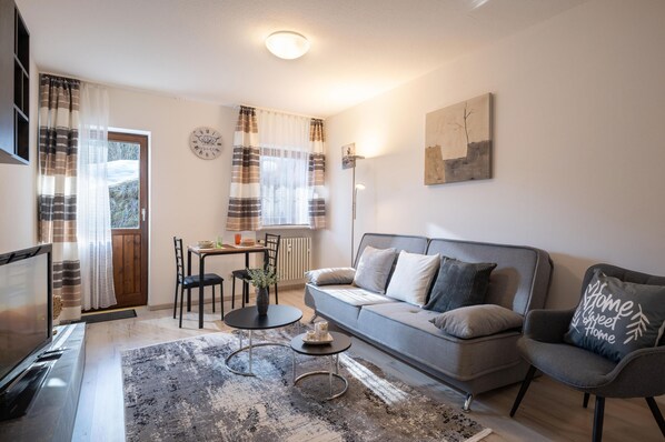 A modern living room with a sofa, armchair, coffee table, and TV. There is a dining table with chairs, a window with curtains, and a wall clock. - 2-room first floor apartment with terrace 4 (Oberaudorf)