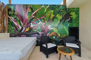 Une pièce comprenant un lit, deux fauteuils en osier noir avec des coussins blancs, une petite table ronde en bois et une fresque représentant une végétation et des oiseaux tropicaux.