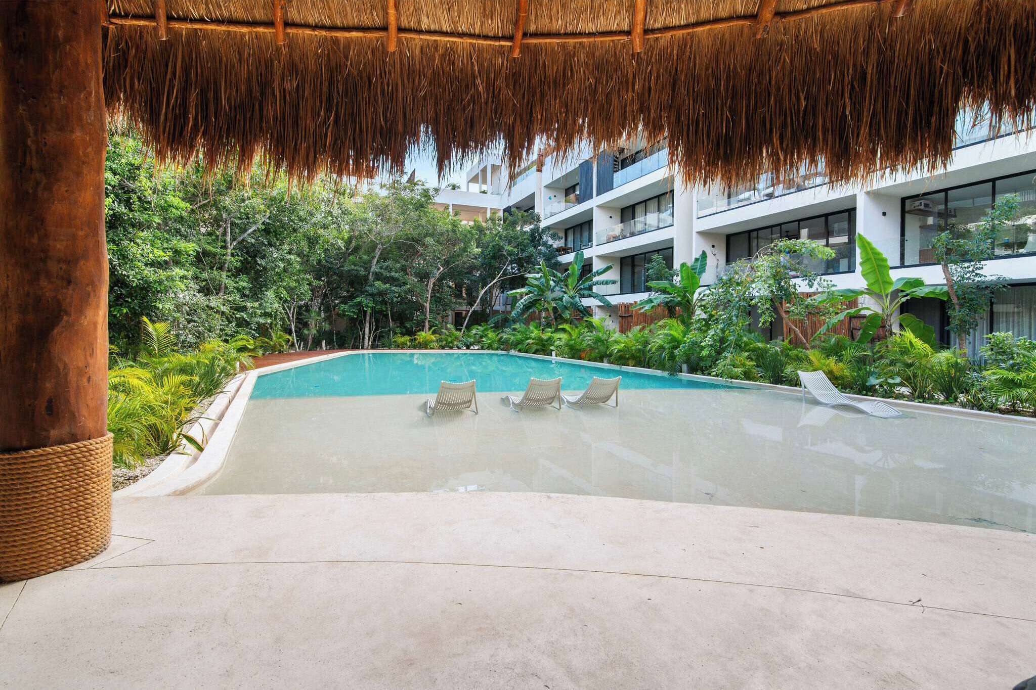 A pool area with lounge chairs, a thatched roof, and lush greenery.