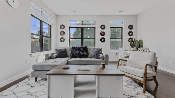Un salon moderne avec un canapé d’angle, une table basse et un fauteuil blanc. De grandes fenêtres sont équipées de stores, un réveil est accroché au mur, et plusieurs disques vinyles sont exposés.