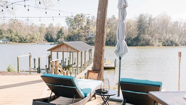 A wooden deck with two lounge chairs and a small table, overlooking a lake with a dock and a wooden structure in the background. - The Marigold at Mill Creek (Topping)