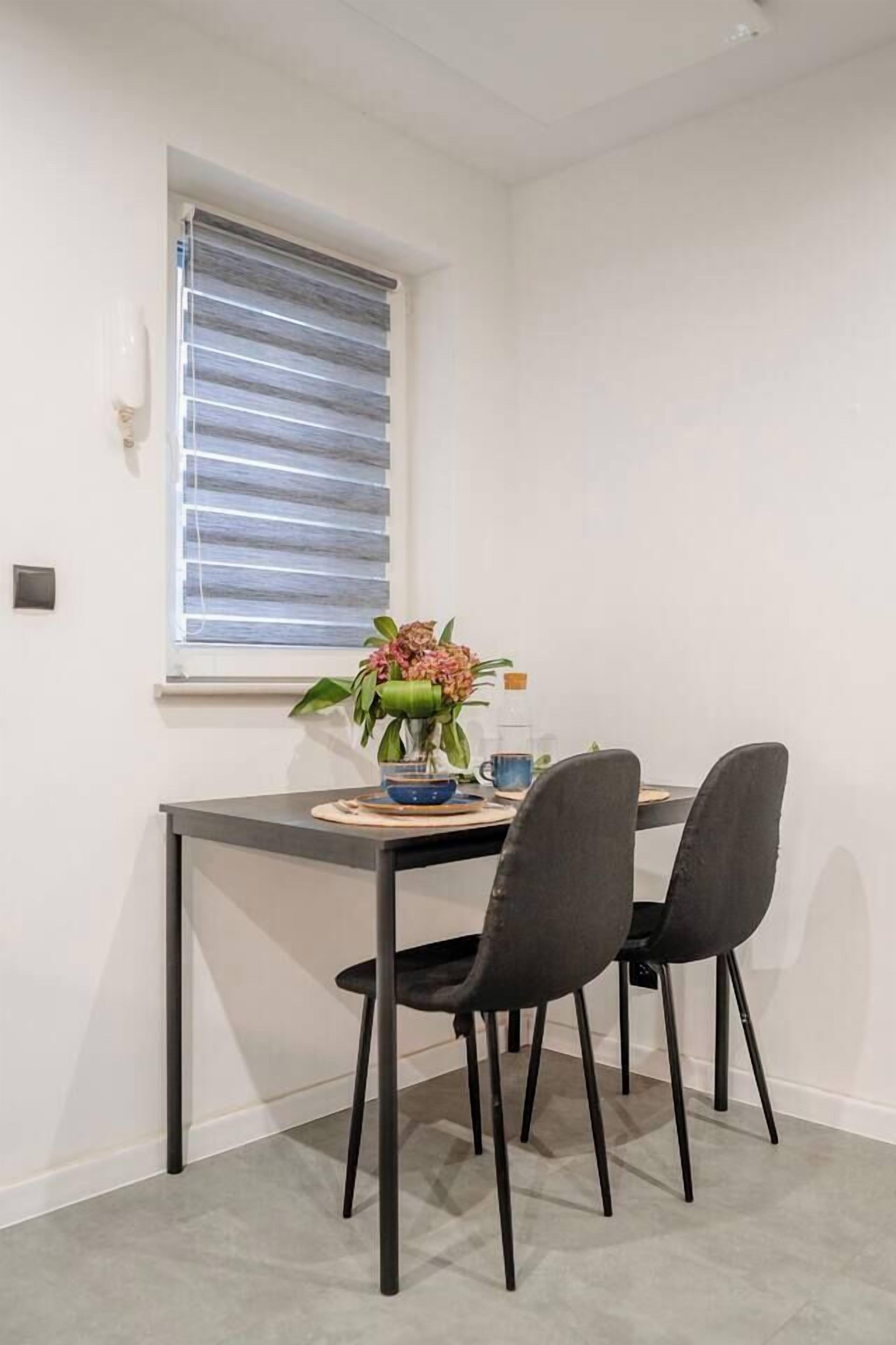 A small dining area with a table, two chairs, a vase with flowers, and a window with blinds.